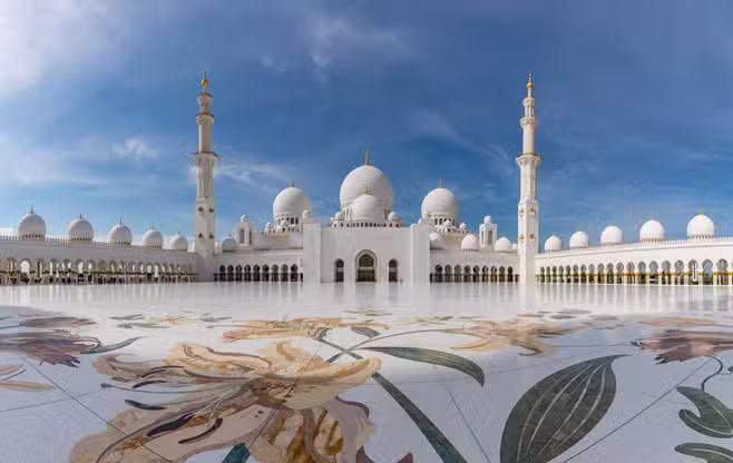 Panoramic view of the Sheikh Zayed Grand Mosque in Abu Dhabi, UAE, showcasing its white marble domes, ornate floral courtyard tiles, and towering minarets under a bright blue sky.