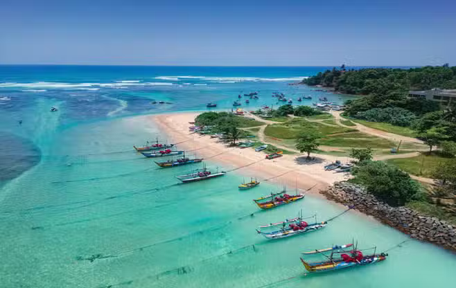 Aerial view of traditional colorful fishing boats anchored in the turquoise waters of Mirissa Beach, Sri Lanka, with a sandy peninsula, lush greenery, and a calm blue ocean under a clear sky.