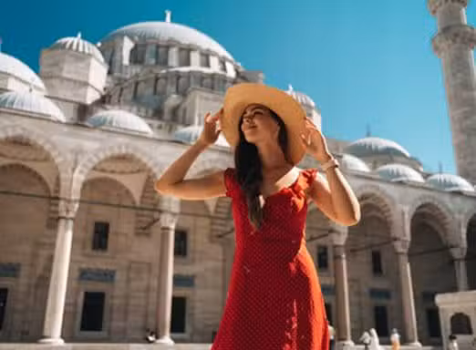 A smiling woman in a red dress and wide-brimmed hat poses in the courtyard of a grand mosque in Istanbul, Turkey, with majestic domes and arches behind her under a clear blue sky.