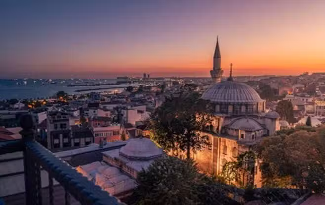 Stunning sunset view over Istanbul, Turkey, featuring a historic mosque with a tall minaret, traditional rooftops, and the Bosphorus Sea dotted with boats in the background.