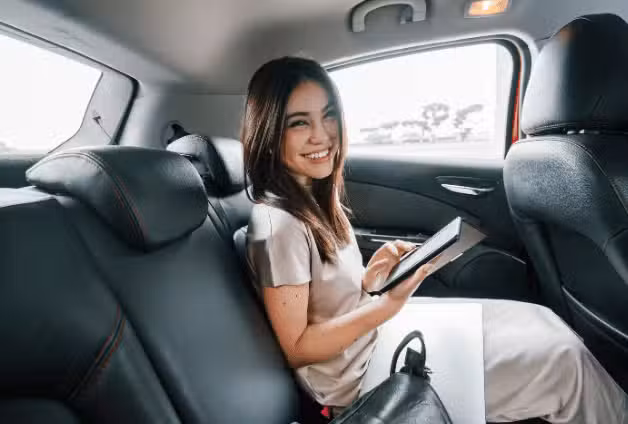 A young Japanese Asian woman sits and reads her e reader while sitting in the backseat of a car she booked via a ride hailing app. She is smiling happily as she reads and is driven to her destination